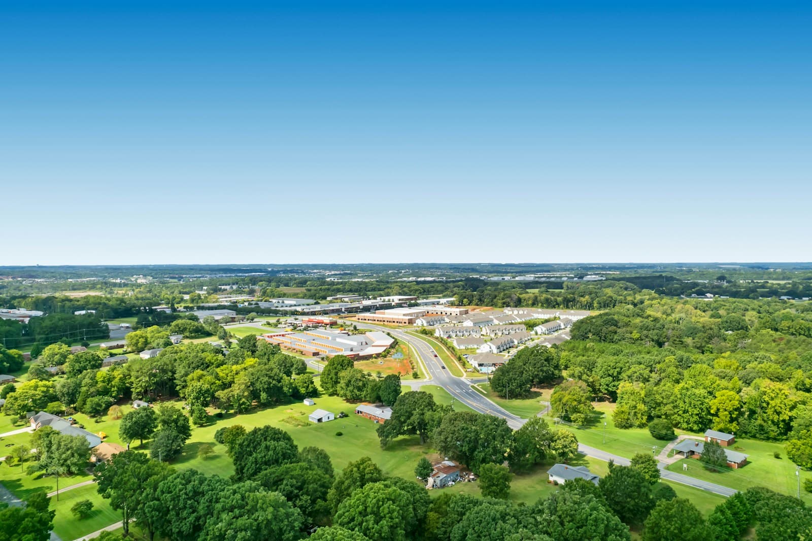 Aerial view of suburban commercial complex surrounded by green trees and open spaces under clear blue sky