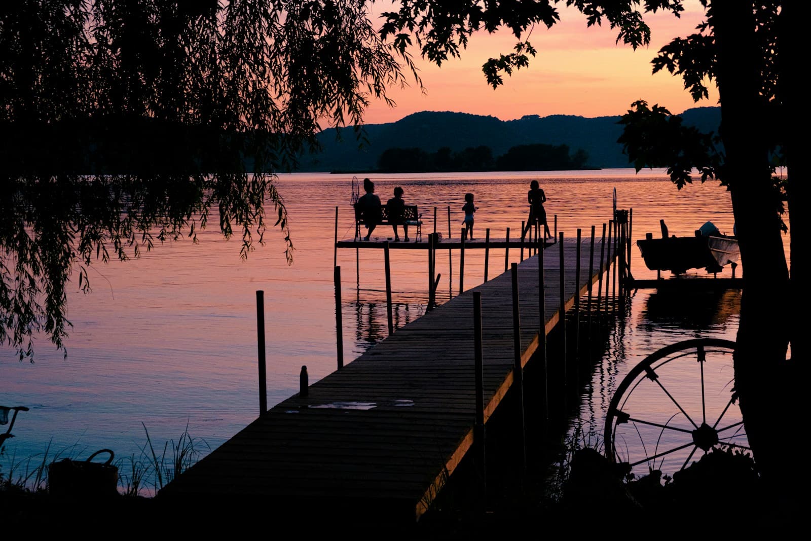 Wooden dock extending into lake at sunset with people fishing, mountains in background, warm golden light