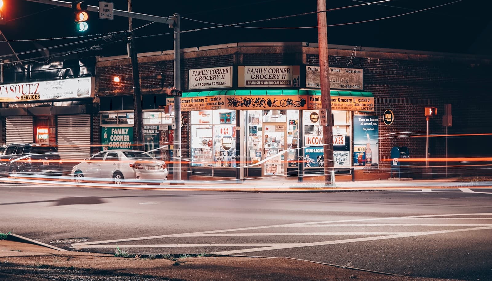 Nighttime street view of illuminated convenience store with light trails from passing cars on wet pavement